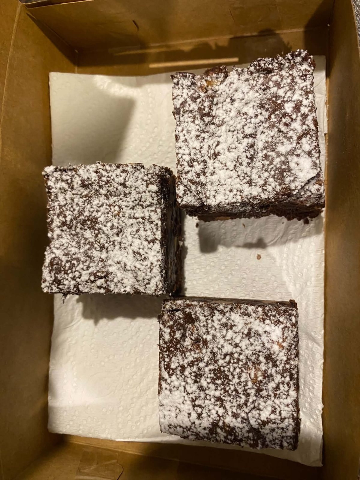 Three thick brownies dusted with icing sugar, nestled in a parchment-lined kraft box.